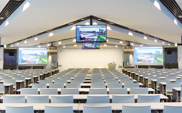写真：追手門学院大学 茨木総持寺キャンパス大教室。ビデオ会議システムによる遠隔授業にも対応し、複数の大型ディスプレイや映像・音響システムが活躍。