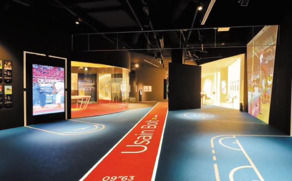 写真：オリンピックミュージアム展示ブースの一部、左に柔道関連の展示、右側に陸上競技に関するブースがあり、それぞれのブースで映像が映し出されている。中央の床にはウサイン・ボルトが実際に出した記録と距離が、陸上競技のトラックを模した赤い枠でシームレスに映し出されている